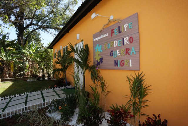 Faixada da casa com parede pintada na cor laranja, jardim com muitas plantas e árvores, letreiro pendurado na parede, porta branca e um caminho até a porta com placas de cimento e grama.