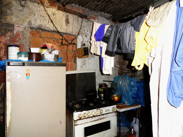 Cozinha de tijolos aparentes, uma geladeira, um fogão, uma mesa com diversos utensílios e um varal de corda com varias roupas.