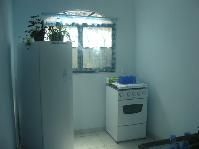 Cozinha com paredes brancas, uma janela com cortina, fogão com copos azuis em cima e geladeira com dois vasos de plantas em cima.