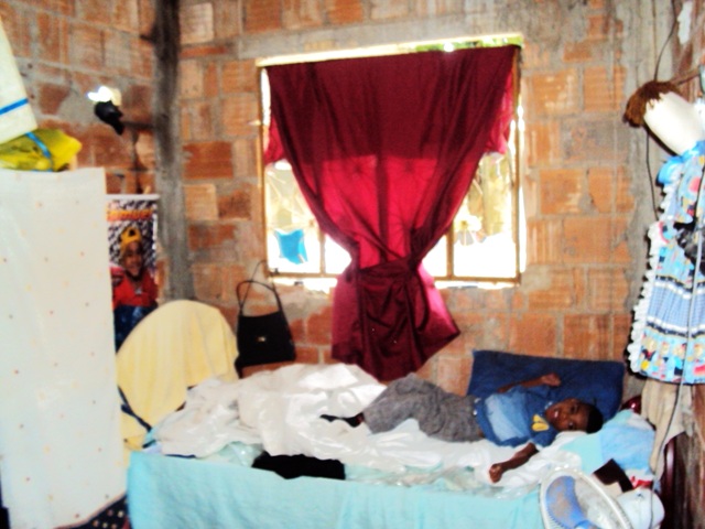 Criança com deficiência na cama do quarto com paredes de tijolos aparentes, janela com cortina vermelha e um ventilador.