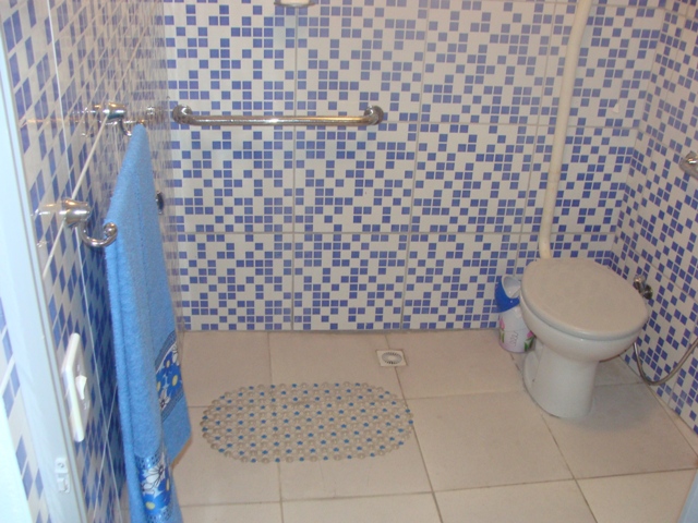 Banheiro com paredes de piso quadriculados em detalhes azuis e brancos, um vaso sanitário, uma barra de apoio de inox, um suporte de toalha com uma toalha azul pendurada e um tapete antiderrapante.