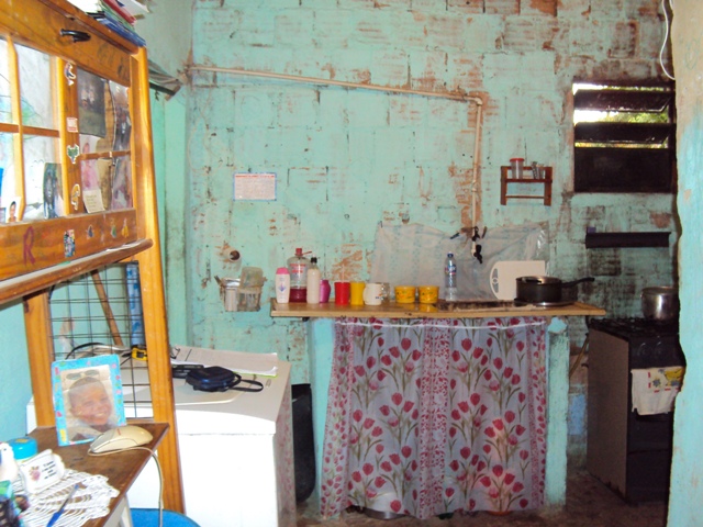 Cozinha com paredes pintadas de verde sobre os tijolos aparentes, um armário, máquina de lavar, fogão e pia com utensílios em cima.