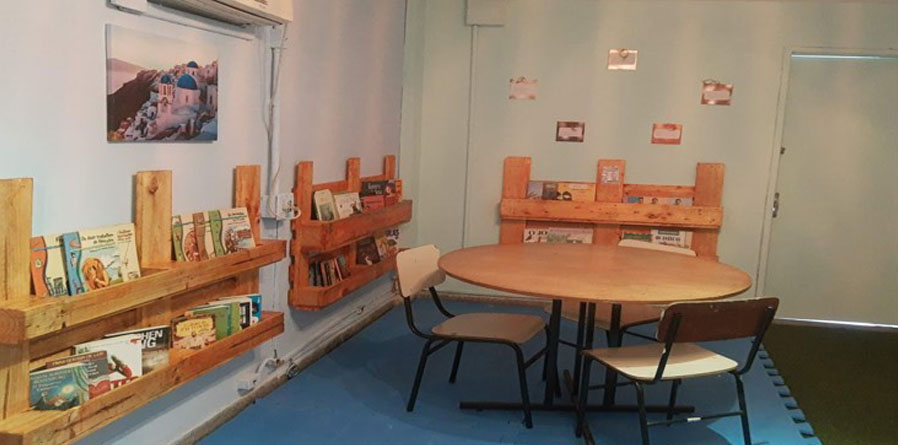 sala com uma mesa redonda ao centro e três cadeiras sob um tatame azul. ao fundo, três prateleiras com livros e seis quadros na parede