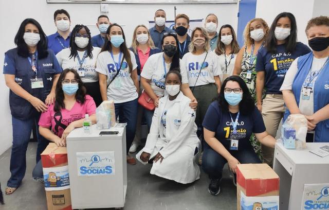 equipes da rioinclui e da clínica da família ocupando uma sala , tendo duas caixas de papelão à frente e balcões de higienização