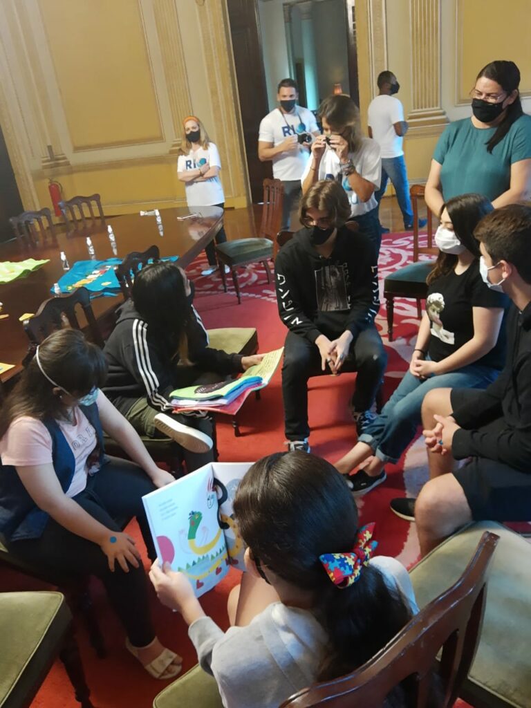 grupo de jovens com e sem deficiência sentados em círculo manuseando livro e conversando tendo adultos observando