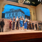 Grupo de artistas no palco do teatro. Parte do grupo na parte baixa do palco e parte do grupo ao longo da escadaria. Ao fundo uma edificação da era colonial.