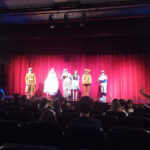 seis adultos num palco usando fantasias. á frente, uma platéia e ao fundo, uma cortina vermelha