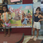 duas meninas sorrindo em torno de um cartaz com o nome da peça teatral