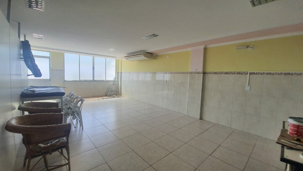 Sala com pisos e azulejos brancos. numa lateral , mobiliário de vime e uma bancada com cadeiras brancas. No fundo, uma janela fechada. No teto, um aparelho de ar condicionado split.