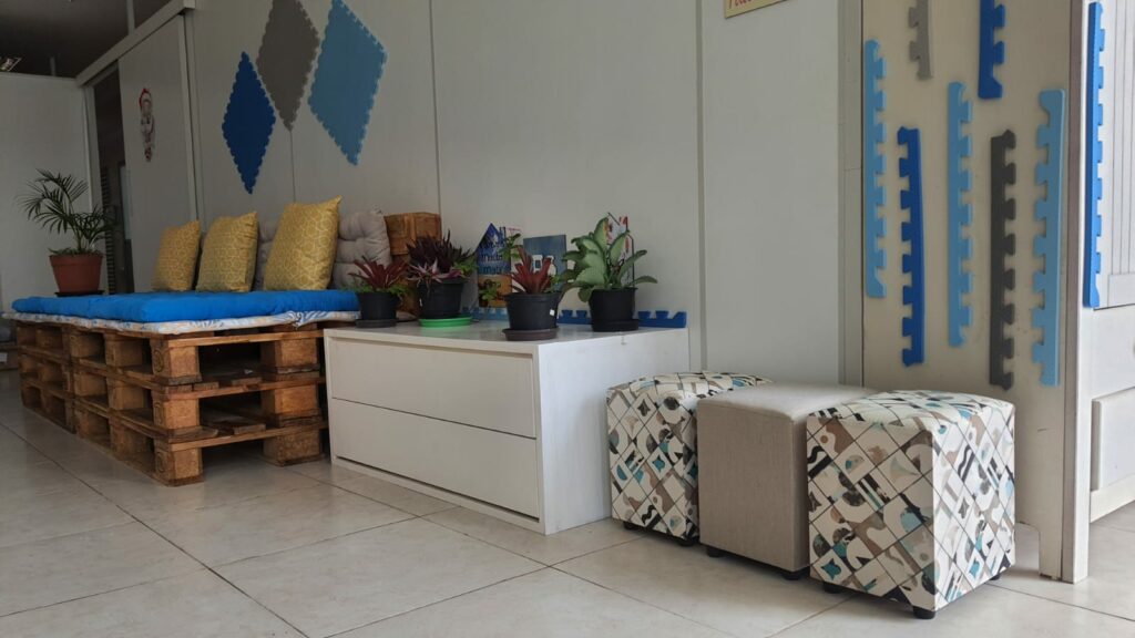 parte de uma sala com sofá feito de palhetes e almofadas azuis e amarelas. Ao lado, gaveteiro branco com vasos de plantas e ao lado, três pufs coloridos