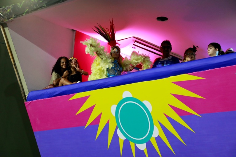 cinco pessoas assistindo desfile de escola de samba no camarote. Ao cento do grupo, uma mulher com alegorias de carnaval.