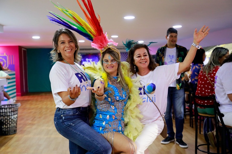 três mulheres sorridentes ao centro da imagem. Uma delas usando fantasia de carnaval