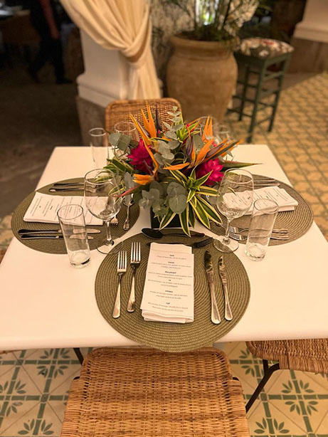 mesa organizada para uma refeição contendo quatro jogos americanos com talheres, cardápio e um arranjo com flores vermelhas ao centro