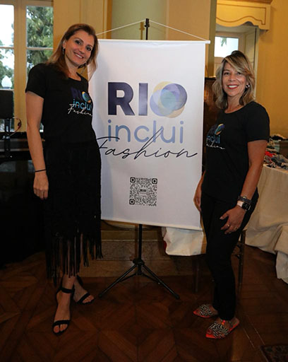 Duas mulheres em pé sorrindo ao lado de um Banner escrito RIOinclui Fashion.