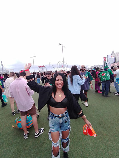 Uma mulher sorrindo com um braço para cima fazendo pose e ao fundo uma multidão no festival de música.