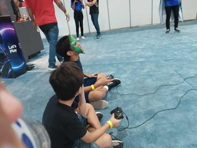 Duas crianças sentadas competindo em jogos de vídeo game, e ao redor pessoas em pé assistindo.