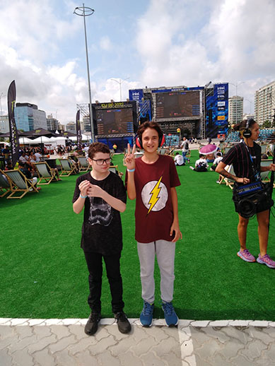 Duas crianças no evento de Game fazendo pose para foto, uma delas com uma camisa com uma imagem de raio, e ao fundo um palco com gramado a frente e pessoas ao redor.