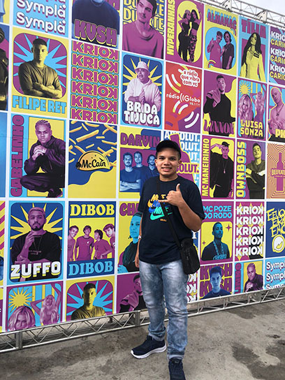 Um jovem fazendo pose para foto, e ao fundo um cartaz com os artistas do festivail.