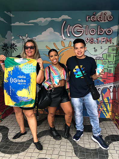 Família com três pessoas em pé sorrindo para foto, com piso parecido com o calçadão de Copacabana e ao fundo uma parede colorida escrito Rádio Globo 98.1 FM