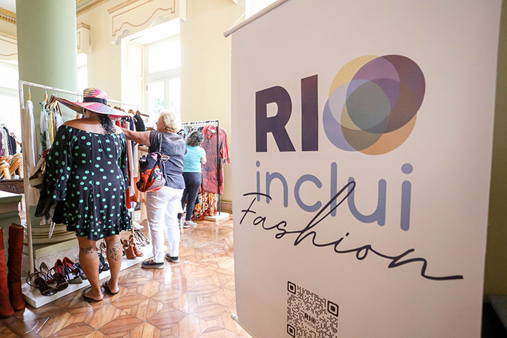 Pessoas escolhendo roupas em bazar realizado no Palácio da Cidade, ao lado o banner do RIOinclui Fashion