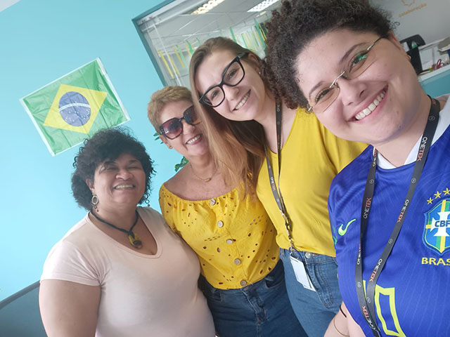 Quatro mulheres sorrindo para foto, e ao fundo uma parede azul com uma bandeira colada do Brasil.