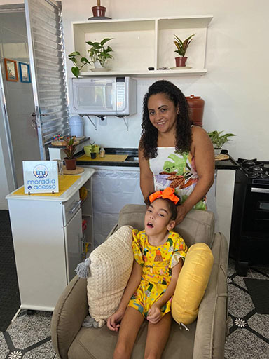 Mãe e filha sorrindo para foto, ao fundo a cozinha reformada pelo Programa Moradia Acesso.