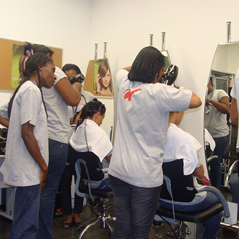 grupo de mulheres de camisetas brancas segurando secadores de cabelo atrás de pessoas sentadas em cadeiras de cabeleireiro em frente a espelhos